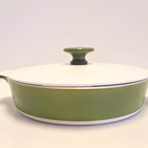 May include: A round, shallow casserole dish with a white lid and a light olive green base. The dish features two integrated side handles and a central knob on the lid, which is also finished in a matching light olive green color.