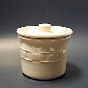 May include: A cream-colored ceramic canister with a matching lid. The exterior features a raised, embossed basket-weave pattern around the middle section. The lid has a small, centered knob handle for lifting. The finish is smooth and glossy.
