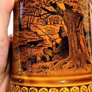 May include: A brown ceramic mug with a silver rim and a black and white illustration of a man fishing under a tree. The mug has a decorative border of small circles.