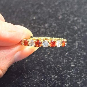 May include: A gold band ring featuring an alternating pattern of round, faceted orange-red stones and clear, colorless stones set in a continuous row around the entire circumference of the metal band, creating a vibrant and symmetrical design.
