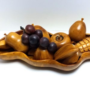 May include: A wooden fruit bowl filled with carved wooden fruits. The bowl contains grapes, pears, a pineapple, and other fruits. The fruits and bowl are a light brown color.