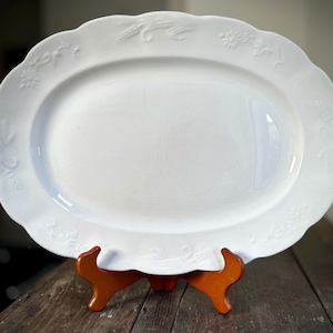May include: A white oval serving platter with a scalloped edge and embossed floral and wheat designs. The platter is displayed on a wooden stand. The overall design is classic and elegant, suitable for serving food.