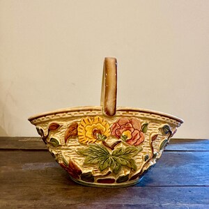 May include: A ceramic basket with a handle, featuring a floral design in shades of yellow, red, and green. The basket has a textured, woven appearance and is set against a neutral background. The handle is a light brown color.