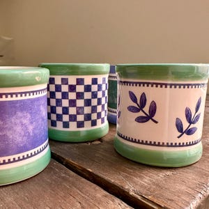 May include: A selection of ceramic pots with green rims and bases. The pots display diverse patterns, such as a chequered design, a watercolour effect, and blue leaf motifs. The pots are placed on a wooden surface.