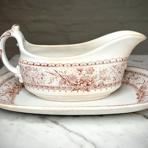 Peut inclure: Saucière crème avec sa soucoupe ovale. L'ensemble présente un motif floral et géométrique de couleur brun rougeâtre. La saucière a une anse et un bec verseur. Un ajout classique à toute table à manger.