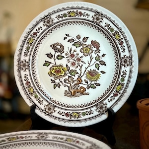 Könnte beinhalten: Ein weißer Keramikteller mit floralem Design. Der Teller zeigt ein zentrales Baum- und Blumenmotiv in Gelb-, Braun- und Rosatönen, umgeben von einem dekorativen Rand. Der Teller wird auf einem schwarzen Ständer präsentiert.
