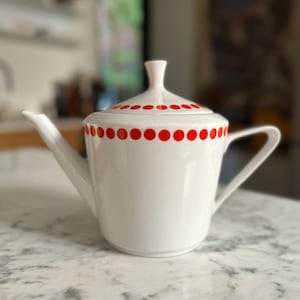 May include: White ceramic teapot with a lid and handle. The teapot is decorated with a row of red polka dots around the body and on the lid. The spout is long and slender. The teapot is sitting on a marble surface.