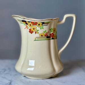 Puede incluir: Jarra de cerámica color crema con un patrón floral decorativo en amarillo, rojo y verde. La jarra tiene un asa curva y un borde festoneado con ribete dorado. El diseño floral se asienta sobre formas geométricas negras y grises.
