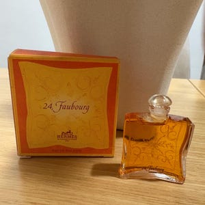Può includere: Un set di bottiglietta di profumo e scatola in miniatura. La bottiglietta di profumo è di forma quadrata trasparente con un tappo di vetro. La scatola è arancione e gialla con il testo "24 Faubourg" e il logo Hermes. Il profumo all'interno è color ambra.