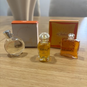 Peut inclure: Trois miniatures de parfum avec leurs boîtes. Les flacons sont transparents et ambrés, avec des bouchons argentés et transparents. Les boîtes sont orange et blanches, avec les noms des parfums imprimés. Les flacons sont disposés sur une surface en bois brun clair.