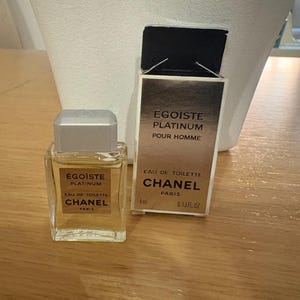 Könnte beinhalten: Ein kleiner Flakon Chanel Egoïste Platinum Eau de Toilette mit silbernem Verschluss und rechteckiger Form. Die daneben stehende Schachtel trägt das gleiche Branding, mit dem Text "POUR HOMME" und "4ml 0.13FL.OZ."