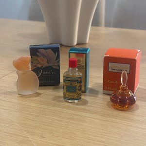 Pode incluir: Três frascos de perfume em miniatura com as suas respetivas caixas. Um é um frasco de vidro fosco com tampa cor de pêssego, um é de vidro transparente com tampa vermelha e rótulo azul, e um é de vidro âmbar com uma tampa transparente.