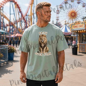 Könnte beinhalten: Hellgrünes T-Shirt mit einem sitzenden Löwen-Motiv und dem Text "FAITH OVER FEAR". Das Shirt besteht aus einem weichen Material und hat eine lockere Passform. Im Hintergrund ist ein Vergnügungspark mit Achterbahn und Riesenrad zu sehen.