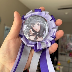 May include: A circular rosette badge featuring a central button with an anime-style illustration of a character with black hair. The rosette is crafted from layered purple, light purple, and silver satin ribbons, with three long ribbon tails hanging down.