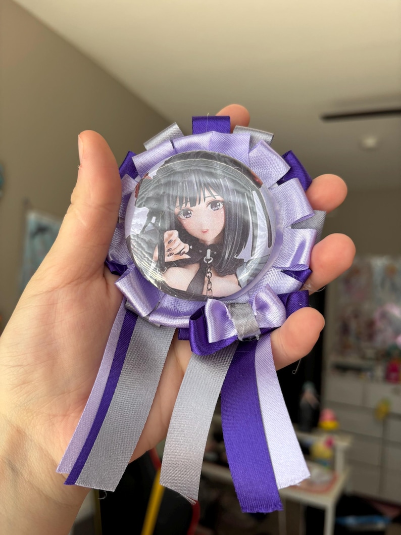 May include: A circular rosette badge featuring a central button with an anime-style illustration of a character with black hair. The rosette is crafted from layered purple, light purple, and silver satin ribbons, with three long ribbon tails hanging down.