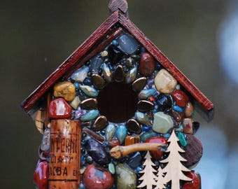 May include: A small wooden birdhouse decorated with a mosaic of colorful stones, glass, and beads. The birdhouse has a red roof and a round opening for birds to enter. The mosaic design includes a sunburst pattern around the opening.