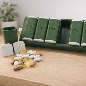 Puede incluir: Un pastillero semanal verde con siete compartimentos etiquetados para cada día de la semana. Incluye un recipiente separado y dos tapas decorativas blancas. Varias pastillas y cápsulas están esparcidas sobre la superficie de madera.