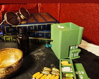 May include: A green plastic pill organizer with slide lock lids labeled with days of the week. The text reads: Three Doses A Day. 169 Colour Combinations. Slide Lock Lids. The organizer contains various white and yellow capsules and tablets.