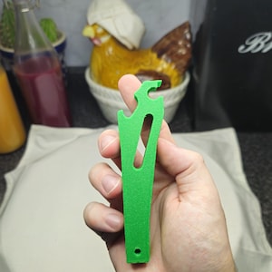 May include: A vibrant green tool with a distinctive design, held in a hand. The tool has a curved top and a central aperture. The background is blurred, showing a decorative chicken ornament and kitchenware.
