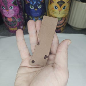 Puede incluir: Un abridor de botellas de metal marrón con un cuerpo rectangular y un pequeño agujero circular. El abridor tiene un borde dentado para hacer palanca. Contenedores decorativos con temática de gatos en el fondo.