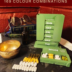 May include: A green plastic weekly pill organizer with a vertical stack of seven labeled compartments for each day of the week. The text SECURE CLICK LOCK LID and 169 COLOUR COMBINATIONS is visible. One compartment labeled MON is open, containing various white and yellow pills.