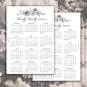 Op de afbeelding: Een witte papieren kalender voor het jaar twintig zevenentwintig met florale illustraties in zwarte inkt aan de bovenkant en een raster voor elke maand. De tekst bevat het jaar twintig zevenentwintig en het label A5 14,8 x 21 cm.