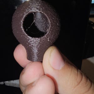 May include: A dark brown, 3D-printed object with a spherical top and a cylindrical base. The sphere has a large central hole. The object is held in a person's hand against a dark background.