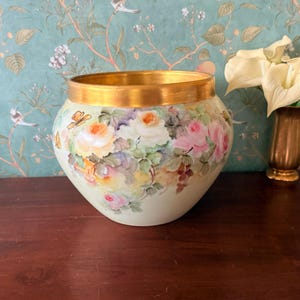 Könnte beinhalten: Eine handbemalte Keramikschale mit goldenem Rand. Die Schale zeigt ein Blumendesign mit Rosen in Rosa-, Gelb- und Weißtönen, umgeben von grünen Blättern. Auf der Seite der Schale ist ein Schmetterling gemalt.