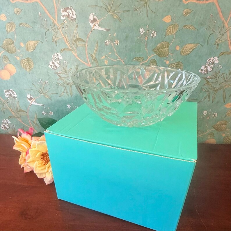May include: A clear, faceted glass bowl sits atop a turquoise box. The bowl has a diamond-like pattern. The background features a floral wallpaper with birds and flowers. A bouquet of yellow and pink flowers is in the lower left corner.