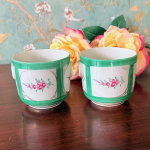 May include: Two white ceramic cups with green trim and handles. Each cup features a floral design with pink flowers and green leaves. The cups are set against a backdrop of flowers and a floral wallpaper.