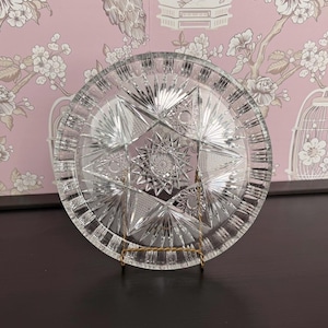 May include: A clear, circular glass plate featuring an intricate, multi-faceted starburst pattern cut into the surface. The design includes radiating lines, geometric triangles, and a central star motif, all supported by a thin, gold-toned wire stand.