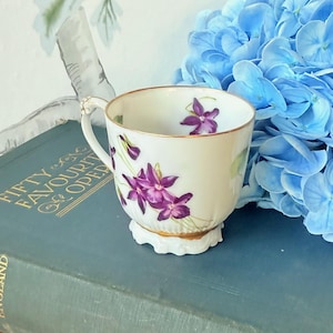 Puede incluir: Una taza de té blanca con borde y asa dorados, adornada con diseños florales morados. La taza se apoya sobre una pequeña base festoneada. La taza de té está colocada sobre un libro azul con letras doradas.