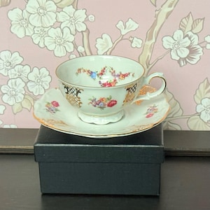 May include: A vintage teacup and saucer set. The cup and saucer are white with gold trim and floral designs in pink, blue, and yellow. The teacup is sitting on a black box.