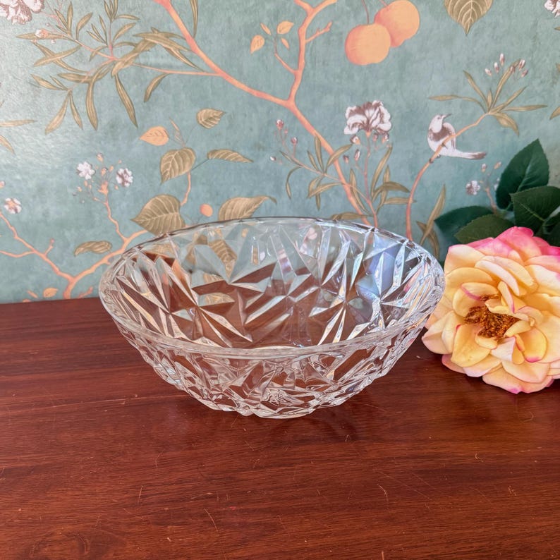 May include: A clear, oval-shaped glass bowl with a geometric, diamond-like pattern. The bowl is displayed on a dark wooden surface, with a floral wallpaper background and a rose in the corner. This decorative bowl is suitable for serving.