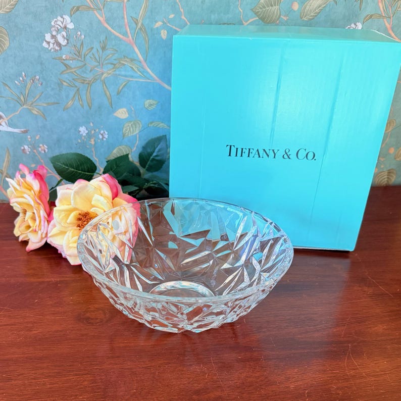 May include: A clear, faceted glass bowl sits on a wooden surface, with a Tiffany & Co. box in the background. The bowl has a geometric design. Pink and yellow roses are to the left of the bowl. The Tiffany & Co. box is a light blue color.