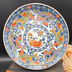 May include: A round ceramic plate featuring a central medallion of orange and blue flowers surrounded by a complex pattern of red, orange, and blue floral motifs, green foliage, and swirling wave designs, all finished with a thin gold rim.