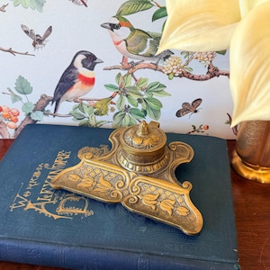 May include: An antique brass inkwell with floral detailing sits atop a dark blue book with gold lettering. The inkwell has a small, lidded ink pot and ornate base. The background features a floral print with birds.