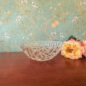 May include: A clear, faceted glass bowl with a geometric design. The bowl is oval-shaped and sits on a dark wooden surface. The background features a floral wallpaper with birds and peach-colored fruit.