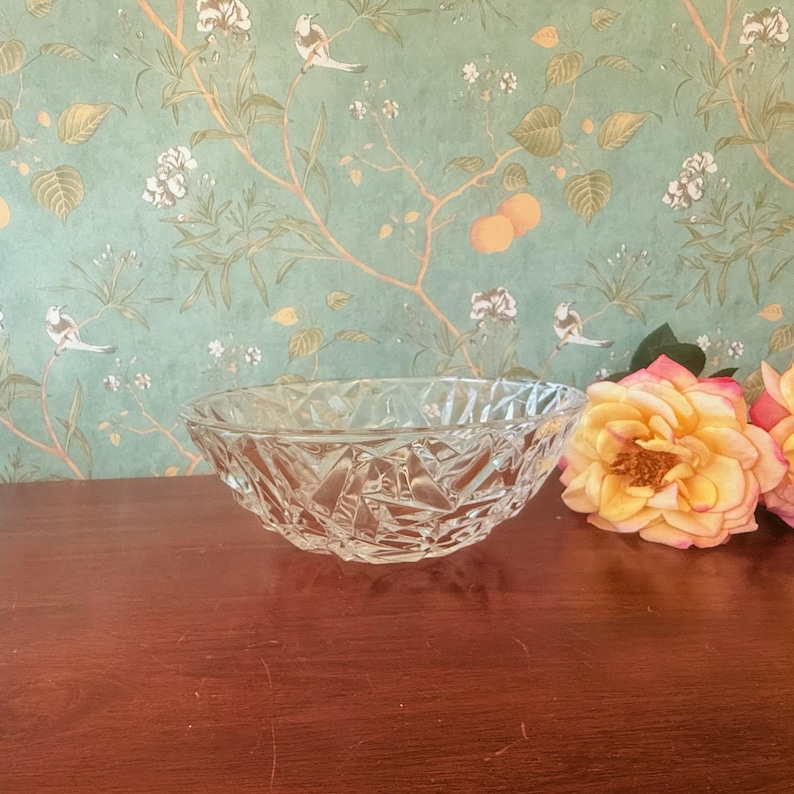 May include: A clear, faceted glass bowl with a geometric design. The bowl is oval-shaped and sits on a dark wooden surface. The background features a floral wallpaper with birds and peach-colored fruit.