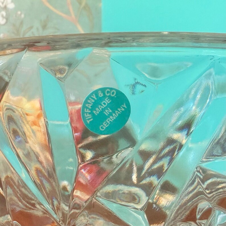 May include: Close-up of a clear glass bowl with a diamond-cut pattern. A round, turquoise sticker with the words "Tiffany & Co. Made in Germany" is affixed to the glass. The bowl reflects light, creating a sparkling effect.