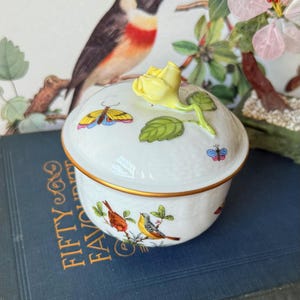 Peut inclure: Une boîte à bijoux en porcelaine blanche avec un couvercle orné d'une rose jaune et de feuilles vertes. La boîte est décorée de papillons et d'oiseaux colorés. Le bord est orné d'or. La boîte est posée sur un livre bleu.
