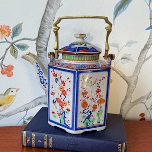 Può includere: Teiera vintage esagonale in ceramica con coperchio e manico in ottone. La teiera è decorata con scene floreali e orientali in rosso, arancione e blu. È appoggiata su un libro blu contro una carta da parati floreale.
