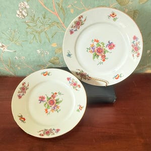 May include: Two white porcelain plates with floral designs. Each plate features a central bouquet of pink, purple, and orange flowers, surrounded by smaller floral clusters. The plates have gold rims and are displayed on a dark wooden surface.
