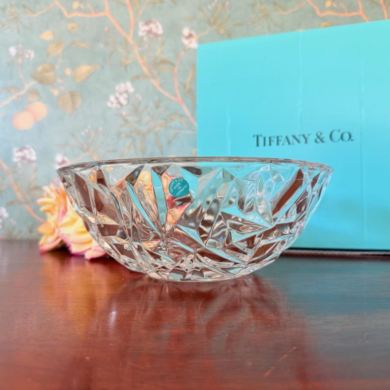 May include: A clear, faceted glass bowl with a diamond-like pattern. The bowl is set against a backdrop of a Tiffany & Co. box and a floral wallpaper. A small, round sticker is visible on the bowl.