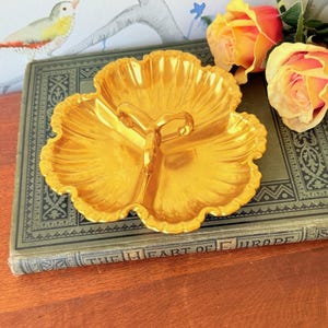 Puede incluir: Una fuente dorada de tres secciones con asa. El plato tiene un diseño en forma de pétalo y está sobre un libro antiguo. Rosas amarillas y naranjas en el fondo.