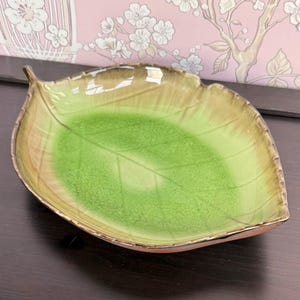May include: A ceramic dish shaped like a leaf with a glossy finish. The center features a vibrant lime green color that transitions into a muted tan or light brown along the edges. The surface has a visible crackle glaze texture and raised veins.
