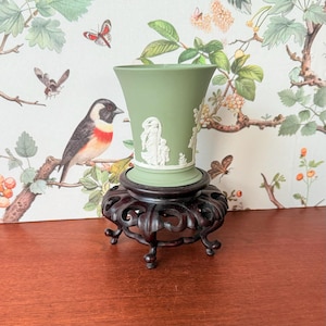 May include: A sage green vase with white relief detailing, set on a dark brown, ornate wooden stand. The vase features classical figures and floral designs. The stand has a carved, openwork design. The background includes a wallpaper with birds and botanical illustrations.