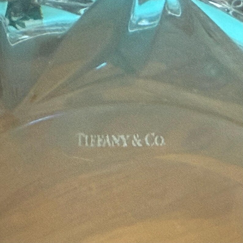 May include: Close-up of a clear, textured glass object with the text "TIFFANY & Co." in white. The glass has a light blue hue and appears to be a decorative item.