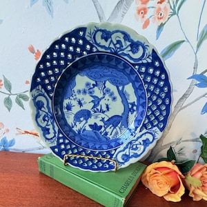 Puede incluir: Un plato decorativo azul y blanco con un borde calado y una escena central de ciervos y pájaros. El plato está sostenido por un soporte de metal dorado, apoyado en un libro verde. El plato tiene un borde festoneado.