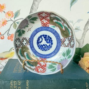 Puede incluir: Un pequeño plato de porcelana antiguo con borde festoneado y un diseño detallado y colorido. El plato presenta un motivo floral azul central, rodeado de patrones geométricos y florales en rojo, verde y dorado. Descansa sobre un soporte de alambre dorado.